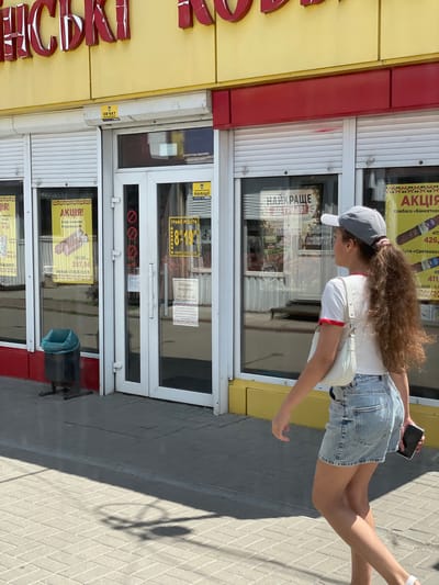 In Cherkasy, Ukraine, a young woman with long, curly brown hair, wearing a grey baseball cap, a white t-shirt with red trim, and a light blue denim skirt, walks past a store. She is carrying a white shoulder bag and a black smartphone in her right hand. The store's facade is yellow with red accents, and it has large glass windows and metal roll-down shutters. Promotional posters for "Акція!" (Sale!) and "НАЙКРАЩЕ ДО ГРИЛЯ!" (Best for grilling!) are visible in the windows. A trash bin with a blue bag is situated outside the store. The ground is paved with light-colored bricks. The image captures a sunny day with strong shadows cast on the pavement.