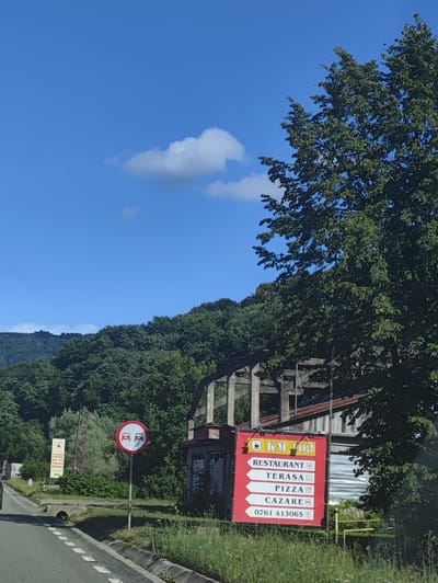 The image captures a roadside scene in Leordina, Romania, under a clear blue sky with a few scattered clouds. On the right side of the frame, a tall tree with dense green foliage partially obscures a weathered, unfinished concrete structure. In front of this structure, a large, bright red sign with white text and arrows serves as a directional marker. The sign reads "KM 108" at the top, followed by "RESTAURANT," "TERASA," "PIZZA," and "CAZARE," with a phone number "0761 413065" at the bottom. To the left of the red sign, a circular traffic sign with a red border and a white background depicts two red cars, indicating no overtaking. Further to the left, a billboard-like sign is visible with text that is partially obscured by foliage. A paved road runs through the foreground, with a white dashed line marking the lane. The surrounding environment is characterized by lush green trees and vegetation, with a hilly landscape in the background. The overall impression is of a rural setting along a road in Leordina.