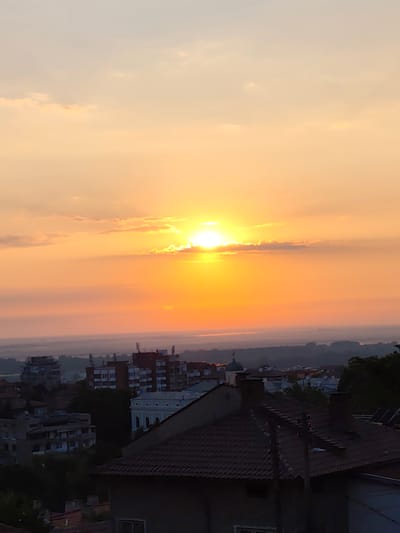 The image captures a sunset over the city of Svishtov, Bulgaria. The sun, partially obscured by clouds, casts a warm, orange glow across the sky, transitioning to a softer yellow and then pale gray towards the top. In the foreground, the rooftops of several buildings are visible, with tiled roofs and chimneys. Some of these appear to be residential homes, while others are larger apartment buildings. Further in the distance, the city skyline is silhouetted against the horizon, with trees and other structures creating a layered effect. The overall scene is serene, depicting the end of the day in an urban setting.