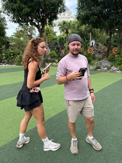 In Hoa Lư Ward, Vietnam, a woman with curly brown hair, wearing a black tank top, a black ruffled skirt, white socks, and grey sneakers, stands with sunglasses on her head. She is holding a white smartphone in her left hand and appears to be smoking. She is looking towards a man standing to her right. The man, with short brown hair and a beard, is wearing a light purple t-shirt, beige shorts, a grey baseball cap, and grey sneakers. He is looking down at a black smartphone in his right hand, which is attached to a red strap. He has a red bracelet on his left wrist and a dark blue and white patterned bracelet on his right wrist. They are standing on a green artificial turf ground with diagonal stripes. In the background, there are lush green trees, palm trees, rocks, and decorative metal flowers. A white building with a blue dome is visible in the far background.