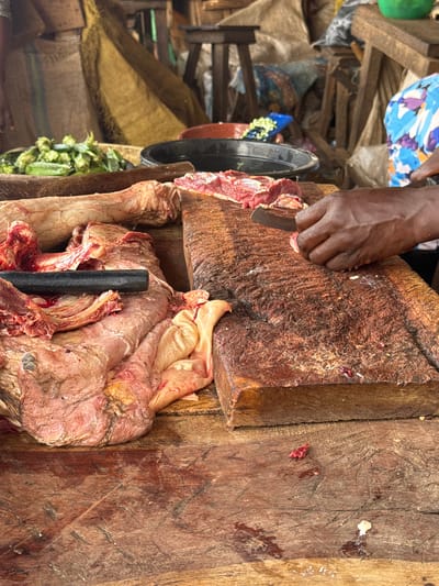 At the Kagwaru market in Nigeria, a butcher's hands are visible as they prepare raw meat on a large wooden chopping block. The butcher, wearing a blue and white patterned garment, is using a curved knife to slice a piece of red meat. The chopping block is worn and stained, showing signs of heavy use. To the left of the block, a large cut of raw meat, possibly beef or mutton, is displayed, with some visceral fat visible. In the background, there are sacks and wooden structures typical of an open-air market. A bowl of green vegetables, possibly okra, is also visible. The scene depicts the preparation of meat for sale in a bustling marketplace environment.
