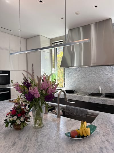 This image depicts a modern, well-lit kitchen located in Naples, United States of America. In the foreground, a large, light grey marble island countertop features a deep single-basin sink with a gooseneck stainless steel faucet. To the left of the faucet, a tall clear ribbed glass vase holds a vibrant arrangement of purple, pink, and white flowers, including stocks and lilies, alongside feathery ornamental grasses. To its left, a smaller, rustic woven basket contains a colorful arrangement of red, white daisy-like, and other mixed flowers. To the right of the sink, a teal leaf-shaped bowl holds several bananas, a red apple, and a yellow lemon. A grey striped kitchen towel hangs over the edge of the sink basin.
The background reveals a sleek kitchen design. Directly behind the sink and island, a series of dark grey or black flat-panel base cabinets are topped with matching light grey marble countertops and a continuous marble slab backsplash extending to the ceiling. Integrated into this countertop is a multi-burner cooktop, with black cast-iron grates visible on two burners, and two stainless steel flush covers over adjacent cooking zones. Above the cooktop and backsplash, a large, brushed stainless steel pyramidal range hood with integrated lighting is prominently centered. To the left of the range hood, white glossy flat-panel upper cabinets extend to the ceiling, with a built-in black oven or microwave visible on the lower left. A tall, narrow vertical window with a view of green foliage and distant structures is positioned between the cabinetry and the range hood. The ceiling is white with several square recessed lights and a long, linear chrome pendant light fixture hangs over the island, providing additional illumination. No people are visible in the scene.
