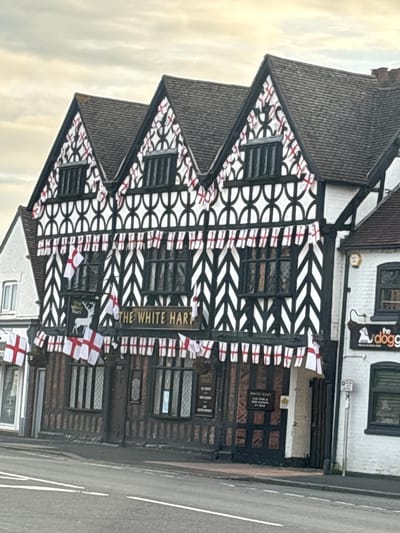 The image shows a three-story historic pub called "The White Hart" in South Staffordshire, United Kingdom. The building features a traditional black and white Tudor-style facade with decorative arches and chevron patterns. The building is adorned with numerous English flags, which are strung along the eaves and windows. The flags are red and white crosses on a white background. The pub has a prominent sign with the name "THE WHITE HART" and an image of a white stag. Adjacent to "The White Hart" is another building, painted white, with a sign that reads "the dogg." The street in front of the buildings is paved and shows white lane markings. The sky is overcast.
