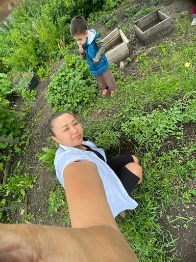 In this vertically oriented selfie, a woman and a young child are positioned in a garden. The woman is on the right side of the frame, taking the photo, with her arm outstretched. She is wearing a white shirt and black shorts, and a hint of her arm and leg are visible. The child, on the left, is wearing a blue puffer vest over a grey hooded jacket and brown pants. He is facing away from the camera, looking down at something he is eating, possibly a berry.

The setting appears to be a garden with rich green foliage and dark soil. In the background, to the left, are two weathered wooden rectangular structures, possibly raised garden beds or compost bins. The overall atmosphere suggests a casual, outdoor activity, possibly a family outing or a moment of observation in nature. The lighting suggests it is daytime, and the lush greenery indicates favorable weather for gardening. The provided location context places the scene in Maloarslangulovo, Russia. There is no visible text in the image.