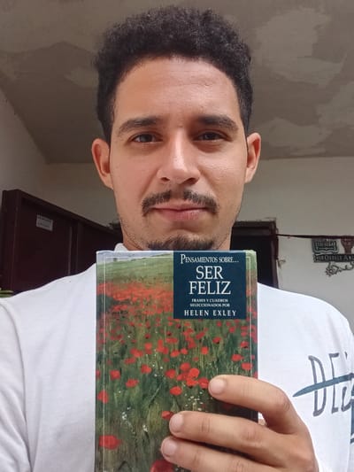 The image shows a man holding a book titled "Pensamientos Sobre... Ser Feliz" (Thoughts About... Being Happy) by Helen Exley. The book has a field of red poppies as a cover image. The man has dark hair, a mustache, and brown eyes. The location is indicated by a sign visible in the background that says "HUELE QUE ALIA ORTEGA AND", indicating that the location is in Duaca, Venezuela.