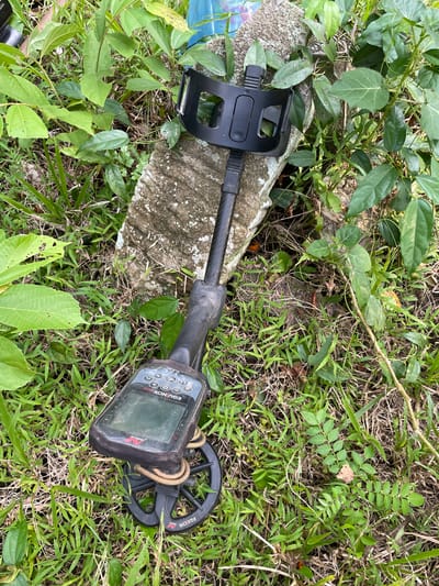 A metal detector lies on the ground amidst lush green vegetation and dry grass, next to a weathered, rough-textured concrete object. The metal detector, a Minelab Equinox 800, is positioned with its coil facing downwards and its control panel displaying readings. The environment appears to be outdoors, possibly a natural area or a less-maintained part of a property, given the density of plant growth. The scene is static, with no people or immediate action depicted. The concrete object suggests a marker or remnant of a structure. The overall impression is of a potentially abandoned or disused location, suitable for metal detecting. The location is identified as Cotseutui, Indonesia.
