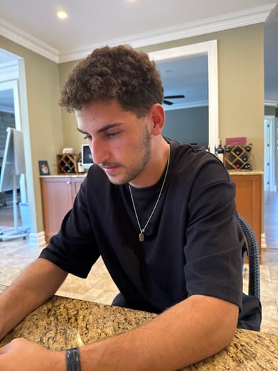 The image captures a young adult male seated at a speckled brown and gold kitchen island in a residential property located in Princeton, United States. The male, observed from the chest up, possesses dark, curly hair and light facial stubble. He wears a dark, short-sleeved t-shirt and a silver chain necklace with a small, rectangular pendant. His attention is directed downwards, suggesting engagement with an unseen object or task on the countertop. A dark wristband is visible on his left wrist. In the background, a domestic interior features light-toned walls, white crown molding, and recessed ceiling lights. A white, mobile easel is partially visible on the left. To the right, a wooden wine rack containing multiple bottles of red wine rests on wooden cabinetry. A doorway in the distance reveals an additional interior space equipped with a ceiling fan. The subject is seated in a chair with a dark, woven back, partially visible on the right.