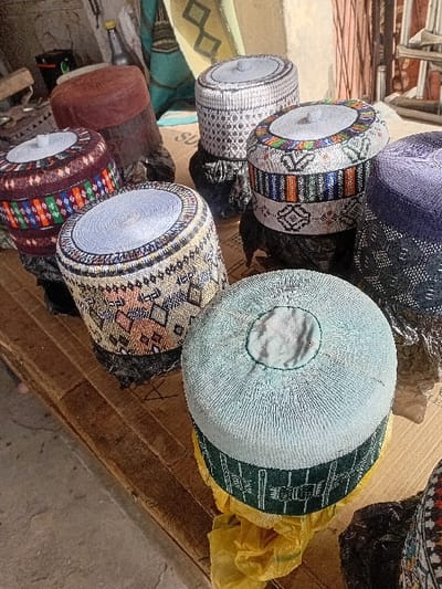 The image captures a detailed arrangement of seven traditional embroidered cylindrical caps, commonly referred to as kufi, displayed on a worn wooden surface in Sharada, Nigeria. Each cap presents distinct hand-stitched patterns and varied color schemes, including shades of white, light blue, maroon, beige, and multi-colored designs. Six of the caps are elevated on small, dark-colored, footed stands, while one light-blue cap rests on crumpled yellow plastic packaging. The background is partially out of focus, revealing a light-colored wall, a section of teal fabric with a diamond motif, an antique cast-iron, a dark bottle, and other indistinct objects. No individuals are present in the scene, and no overt actions are depicted.