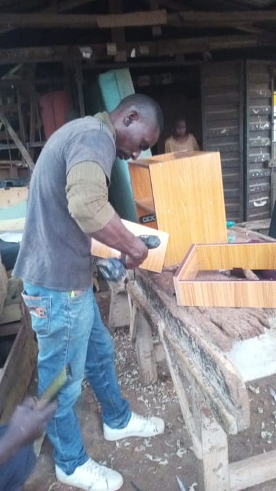 In Barikin Sabon Gidan Kanat, Nigeria, a man is working on a wooden furniture piece in an outdoor workshop. He is wearing a gray t-shirt, a light brown sweater or undershirt, and blue jeans. He is holding a piece of wood and a tool, possibly a sander, in his hands, and appears to be in the process of sanding or assembling the wood. Another person, partially visible, is in the background, inside a building. The workshop environment is rustic, with visible wooden structures and tools scattered around, suggesting ongoing carpentry work. The ground is dirt, with wood shavings present.