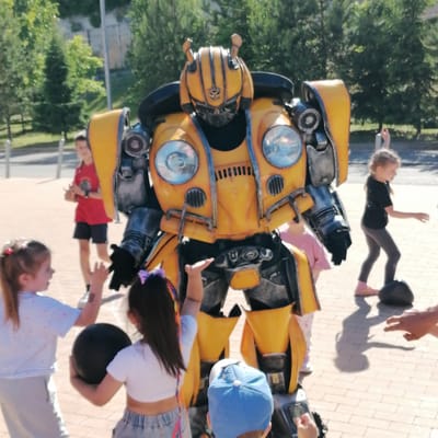 A large, yellow and grey costume resembling the Transformer character Bumblebee stands centrally on a paved outdoor surface, facing slightly left. The costume features circular headlights on its chest, a prominent Autobot symbol on its helmet, and antenna-like structures on top. Several children are gathered around it.
In the left foreground, a young girl with light hair, wearing a striped white top and grey pants, looks up at the costume with her left hand raised. She holds a black ball in her right hand. To her right, another young girl with dark hair, wearing a white crop top and denim shorts, also raises her left hand towards the costume while holding a black ball in her right. Partially visible at the bottom, a young boy in a blue baseball cap looks upwards.
To the left of the costume, a boy in a red t-shirt and black shorts stands, observing the scene. To the right of the costume, a girl in a black t-shirt and dark leggings stands with her arms slightly raised, with a black ball near her feet. A partial adult hand is visible in the lower right, pointing towards the girl and the ball.
The ground is paved with light-colored tiles, exhibiting distinct shadows from the figures, indicating bright daylight. The background features lush green trees and foliage, characteristic of an urban park or public garden in the city of Трубачеевка, Russia.