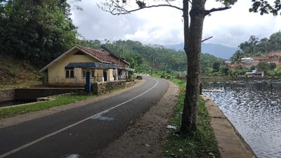 This image depicts a serene, rural landscape in Kecamatan Panjalu, Indonesia, under an overcast sky during the daytime.

A paved asphalt road, featuring a clear white center line, curves from the foreground towards the middle ground, inviting exploration into the hilly surroundings. On the left side of the road, a simple, single-story house with light yellow walls and a reddish-brown tiled roof is prominent. It has a blue-accented porch area and sits adjacent to a narrow, concrete-lined canal or ditch. Behind this house, a lush, verdant hillside rises steeply, densely covered with trees and tropical vegetation.

To the right of the road, a calm body of water, likely a lake or a large pond, extends into the distance. The water's edge is defined by a concrete path, and within the water, multiple structures resembling fish pens or aquaculture nets are visible, suggesting local livelihood activities. A large tree trunk stands in the right foreground, its branches extending overhead.

In the background, beyond the water and the immediate hills, more houses with reddish roofs are nestled among the trees, dotting the landscape. Rolling, tree-covered hills form the distant horizon. The scene evokes a sense of peaceful rural life, characterized by natural beauty and human settlement harmoniously integrated with the environment. No specific activity or event is taking place, and no text is visible.