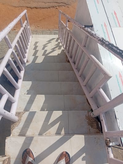 The image shows an outdoor staircase with light pink metal railings. The stairs are made of light-colored tiles and are partially covered in shadow from an unseen object. The area around the stairs is dusty and appears to be a construction site, with loose gravel and dirt visible. On the right side of the image, a metallic surface with text and red arrows is attached to the wall next to the railing. The visible text on this surface, in English, includes instructions like "PLEASE TEAR OFF PROTECTIVE FILM" and "45DAYS AFTER INSTALLATION". At the bottom of the image, two feet wearing flip-flops are visible, suggesting someone is standing on the stairs. The lighting indicates it is daytime, and the shadows suggest a sunny day. The overall scene depicts an unfinished or recently constructed outdoor area, likely in a warm climate given the dusty ground and outdoor setting.
