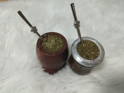 The precise location in the city of al Burj, Palestinian Territory, cannot be determined from the visual information provided in the image.
The image displays two yerba mate gourds (mates) with accompanying bombillas, positioned on a white, fluffy surface resembling faux fur or a soft blanket. The gourd on the left is dark reddish-brown, appearing to be made of wood or a similar natural material, with a smooth, slightly tapered cylindrical shape. The gourd on the right features a silver-colored metal rim with embossed or engraved patterns, resting atop a darker base that appears to be wood or a natural gourd. Both gourds are filled to the brim with green, dried yerba mate leaves. Each gourd contains a silver-colored metal bombilla (filtered straw) inserted into the mate, with a decorative four-petal design visible near the top of each straw. No individuals or actions are depicted; the objects are static.