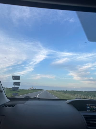 The image shows a view from inside a car driving down a road toward a distant cluster of buildings that appear to be historical structures. The road is paved and has white lines marking the lanes. On either side of the road, there are fields of green grass and some crops. The sky is blue with scattered white clouds. In the lower right corner of the image, a dashboard display shows the temperature is 23 degrees Celsius, average fuel consumption is 33.0 MPG, and the speed is 150. The buildings in the distance are likely the Chateau de Sacy, located in Sacy-le-Grand, France, given the rural landscape and architectural style. There are no people visible in the image, and no animals or other objects of note apart from the car's interior and dashboard.