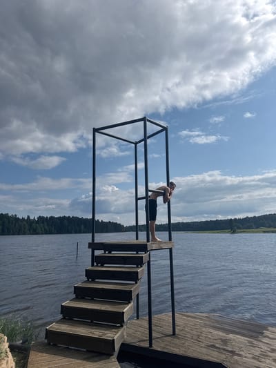 A young man stands on a wooden dock overlooking a lake in Pozdyshki, Russia. He is wearing black swim trunks and has his arms crossed over his chest, looking upwards. The dock is made of weathered wood planks and has a metal railing. Steps lead down from the dock into the water. The lake is calm and reflects the cloudy sky. A dense forest lines the opposite shore of the lake. The time of day appears to be afternoon, with the sun partially obscured by clouds. The overall mood is peaceful and relaxed, suggesting a day of leisure by the water.