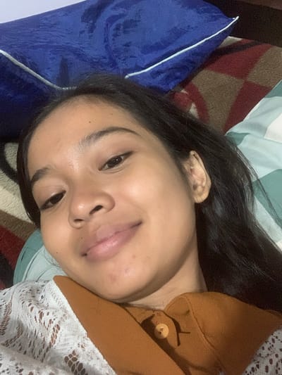 A young woman with dark hair and brown eyes lies on her back, looking at the camera with a slight smile. She is wearing a white lace top and a rust-colored button-up shirt. The background consists of a blue pillow and a patterned blanket. The lighting suggests an indoor setting, possibly a bedroom. The image was captured in Sungai Meriam, Indonesia.
