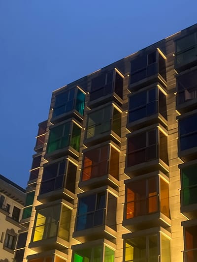 This is a low-angle, medium shot of the exterior of a modern building at dusk in Bilbao, Spain. The building features multiple staggered balconies with colored glass panels, illuminated by warm, yellow lights. The sky above is a deep, clear blue. To the left of the main building, a portion of an older, lighter-colored building with arched windows and wrought-iron balconies is visible. The main building's balconies are recessed and appear to be constructed of concrete or a similar material, with thin yellow LED strip lights outlining their edges. The colored glass panels are in shades of blue, green, yellow, and orange, reflecting the interior lights and the twilight sky. The overall impression is one of contemporary architecture against a darkening sky.