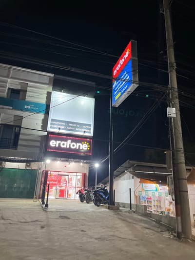 This media file depicts a nighttime scene outside a storefront in Bundar, Indonesia. The primary focus is on illuminated signage advertising "erafone," a telecommunications or electronics retailer. Multiple signs are visible, including a prominent red and blue rectangular sign with the "erafone" logo and text mentioning "HANDPHONE," "SMART TV," "LAPTOP," "SMARTHOME," and "AUDIO ACCESSORIES." Another sign, a long horizontal red one with the "erafone" logo in white, is also clearly visible.
The storefront itself appears to be modern, with large glass windows revealing shelves stocked with various products, predominantly in red packaging. In front of the store, several motorcycles are parked. The overall setting suggests a commercial area, likely a street with pedestrian and vehicle traffic, though no people are prominently featured in the foreground. The lighting indicates it is nighttime, and the concrete ground is visible, suggesting an outdoor urban environment. The image captures the vibrant, illuminated facade of the business as part of its nighttime operations. The presence of multiple wires and poles suggests overhead electrical infrastructure, common in many urban settings.