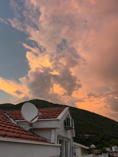 The image shows a sunset sky with dramatic clouds illuminated in shades of orange, pink, and yellow. A densely forested hillside dominates the right side of the frame, with a few buildings visible near the top. In the foreground, the terracotta tiled roof of a building is visible, featuring a satellite dish and an air conditioning unit. The orientation of the image is on its side, with the sky occupying the upper left portion and the hillside and buildings on the right. The scene appears to be a residential area in a hilly, coastal location, possibly during a calm evening. The soft, warm light of the sunset and the vast expanse of colorful clouds create a serene and picturesque atmosphere. No people are visible in the image. The satellite dish has "m'SAT" printed on it.