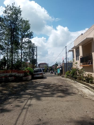 The image depicts a dirt road in a residential area. On the left, there are tall trees and a red and white fence. A silver van is parked on the side of the road. In the distance, several people are walking away. On the right, there is a tan-colored building with potted plants along the side. Power lines stretch across the sky, which is blue with white clouds. The location appears to be a neighborhood in Zeyathiri District, Myanmar.
