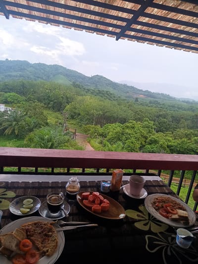 This is an outdoor scene on a balcony. There is a table with breakfast items including bread, omelets, tomatoes, papaya, coffee, cucumber, and yogurt. The balcony has a reddish-brown railing with a mountain and forest landscape in the background. The location is likely Ban Tak Daet, Thailand. The sky is overcast. The balcony has a roof made of wood and dark metal beams.