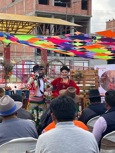 The image captures a cultural event in the city of El Alto, Bolivia. A woman in a red traditional dress and a man in a black hat and colorful traditional attire are performing or presenting. They stand on a decorated stage with colorful streamers overhead. The crowd surrounding them is mostly men with traditional hats on. The background contains buildings under construction and a banner for "CONEXION".