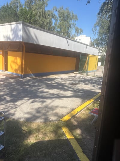 The image depicts an outdoor scene adjacent to a yellow building with a white roof. The building appears to be a facility with open windows and a shaded entryway. In front of the building, there is a paved area with gravel and scattered shadows from trees. To the right, a green metal fence encloses a yellow structure. In the foreground, a yellow and black striped speed bump is visible, and further to the right, a small red and blue folding chair is set up on the grass. The sky is clear and blue, with trees visible in the background. The location is identified as Gebersdorf, Austria. There are no people or active interactions visible in the image.