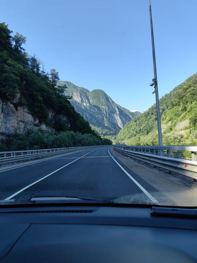 The image captures a view from inside a vehicle driving on a paved road through a mountainous, forested region. The road has two lanes in the direction of travel, with white lane markings. A metal guardrail runs along the right side of the road. On the left, a steep, rocky cliff face is covered with dense green foliage. On the right, a similar steep, forested mountainside rises. A tall pole with surveillance equipment is visible on the right side of the road. The sky is clear and blue. The scene appears to be an outdoor landscape with natural scenery and infrastructure for transportation. The specific location in the city of Monastyr, Russia is not identifiable from the visual information in the image.