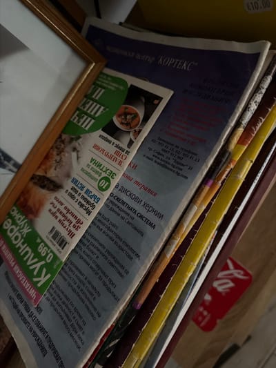The image depicts a close-up, slightly angled view of several stacked magazines and brochures, primarily in Bulgarian, resting against a wooden frame. The lighting suggests an indoor environment, possibly a home or office, with natural light casting shadows.
The top magazine, a blue and white publication, displays the text "МЕДИЦИНСКИ ЦЕНТЪР "КОРТЕКС"". Beneath this, there is a list of services including "ПРЕПОРЪЧВАНИ КОМОГРАФИИ ИЗСЛЕДВАНИЯ". Further down, text details "Записване на телефон:" followed by a phone number and website address "www.kortex-bg.com". A green brochure is visible underneath, featuring text such as "10 ИНТЕРЕСНИ ФАКТА" and "ЗЕЛЕНЧУКОВИ".
Another magazine has a prominent green section with a price of "0,99" and the word "КУХНЯ". There are also sections with text like "АПАЛУДИ" and "ЗДРАВЕ". The overall impression is one of a collection of health, wellness, and culinary publications.
There is no visible indication of the city of Dromolaxia - Meneou Municipality, Cyprus within the image itself.