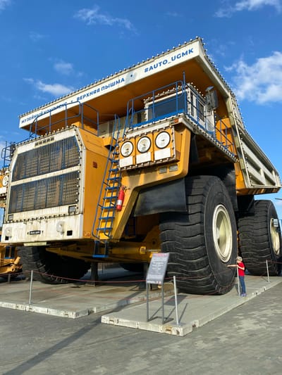 This is a photograph taken outdoors under a blue sky with scattered clouds. The main subject is a very large, yellow mining truck, likely a BelAZ, displayed as part of a museum complex. The truck is predominantly yellow with white and blue accents. It has massive black tires. Blue railings and stairs are visible on the side of the truck, leading up to the cab. A red fire extinguisher is attached to the stairs. The words "МУЗЕЙНЫЙ КОМПЛЕКС" and "ВЕРХНЯЯ ПЫШМА" (Museum Complex Verkhnyaya Pyshma) are written in blue text along the top edge of the truck. A hashtag "#AUTO_UGMK" is also visible in blue text.

In the foreground, a small child wearing a red shirt, blue jeans, and a patterned cap stands near the base of the truck, interacting with a display. The child is on a raised platform that is roped off with red cord. A metal information board is also present on the platform. The ground is paved with gray cobblestones. The overall scene suggests an outdoor exhibition or museum in Verkhnyaya Pyshma, Russia, showcasing large mining equipment.