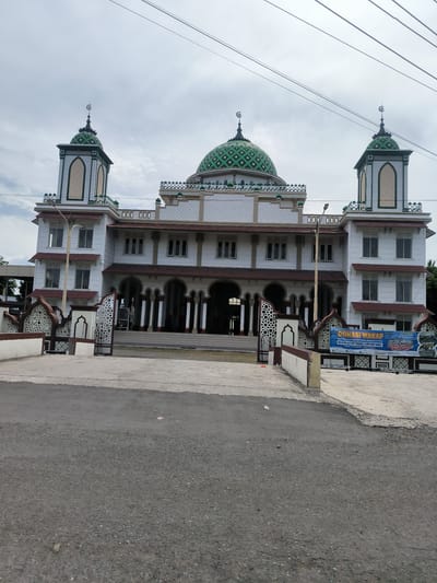 A wide-angle, eye-level outdoor shot captures the Grand Mosque Tengku Lampuk Saka in Dua Paya, Aceh, Indonesia, on an overcast day. The mosque features a prominent white facade adorned with green and white decorative patterns. A large central dome and two smaller domes atop minarets are covered in a distinctive green and white checkered design, each capped with a crescent moon and star finial. The building displays multiple arched windows and entrances, with brown-tiled roofs on its lower sections.
In the foreground, an asphalt road transitions into a concrete pathway leading to two ornate metal gates, which are integrated into a decorative fence featuring intricate white and brown Islamic-inspired patterns. A tall streetlamp stands to the left of the main entrance. Overhead, several utility wires crisscross the cloudy sky.
No people or specific activities are visible, presenting a serene architectural view. On the right side, a blue and white banner provides details for "DONASI WAKAF" (Waqf Donation) "UNTUK PEMBANGUNAN & RENOVASI RUMAH IMAM" (for the construction and renovation of the Imam's house) for "MASJID RAYA TENGKU LAMPUK SAKA." The banner also specifies its location as "JL. RAYA BANDA ACEH MEDAN KM. 13 GAMPONG DUA PAYA KEC. INGIN JAYA KAB. ACEH BESAR," confirming the geographic context. Banking details for "BANK ACEH" and contact phone numbers are also displayed on the banner.