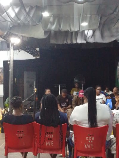 The image depicts an indoor gathering, possibly a performance or presentation, with an audience seated on red chairs facing a stage area. The ceiling features decorative fabric drapes illuminated by spotlights. In the foreground, two individuals are seated on red chairs, their backs to the camera. The chair on the left has the number "005" marked on its back, while the chair on the right has "013 WE" marked.
Beyond the foreground, several other individuals are visible, some seated and some standing. Some are looking towards the front, suggesting they are observing something on the stage. The background is dominated by a dark curtain. The scene appears to be from a public event or venue. The presence of the inscription "WE" on a chair, in conjunction with the general atmosphere, might suggest a theme or a collective group involved in the event.
This image is located in Osogbo, Nigeria, likely at a venue designed for performances or gatherings. The setup with seating and a stage area is characteristic of such locations. The lighting and the presence of an audience indicate a planned event taking place.