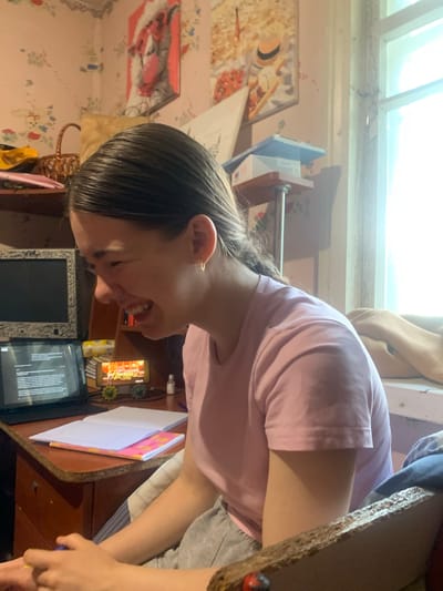 A young woman with dark hair pulled back is sitting at a desk in a room. She is wearing a pink t-shirt and grey sweatpants. She is laughing and looking to her left. On the desk are a laptop, a tablet displaying text, an open notebook, a pink notebook, and a small toy train with a banner that reads "ПАЦАНКИ НАВИГАЦИЯ". The room has pink floral wallpaper and a window with white trim. A basket and a painting of a cow wearing a Santa hat are visible in the background. The scene appears to be indoors, possibly in a residential setting in Hoholeve, Ukraine.