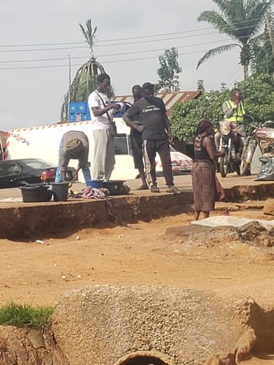 In Kachia, Nigeria, a group of individuals are gathered near a road. In the foreground, a person is bent over, appearing to wash or handle items in large black containers situated on a raised dirt embankment. Next to these containers are folded clothes. Behind this activity, a white van with red and blue markings is visible, parked on the road. Several men are standing around the van, one of whom is holding a white object. Further down the road, a man wearing a reflective yellow vest is seated on a motorcycle. Another motorcycle is parked nearby, and a woman wearing a patterned skirt and a dark top is standing next to it. The environment appears to be a street or market area, with trees and buildings visible in the background. The ground is primarily made of dirt and sand.