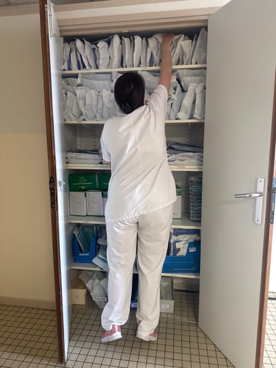 A person wearing white scrubs and pink sneakers is lying horizontally inside a cabinet or closet with open doors. The cabinet shelves contain various medical supplies, including individually wrapped items, boxes of sterile gauze compresses, and boxes labeled "Wuhrlin-Soplamed." The location is indicated by the text on the box and the company name on the box. The boxes say "Wuhrlin-Soplamed" which indicates that this image was taken at their headquarters in Tourcoing, France. The background shows a tiled wall.