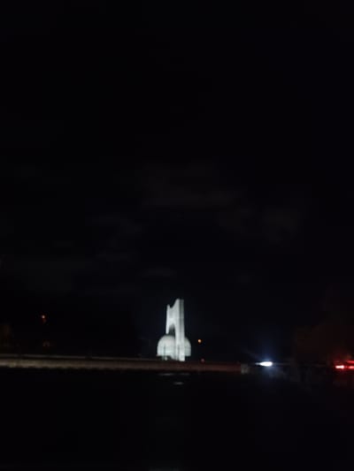 A nighttime shot reveals a distant, illuminated building with a distinctive, layered structure. A tall, slender pole, potentially a tower or antenna, stands prominently in the foreground, stretching upwards with a light at its peak and a red marker further up. The surroundings are dark, with minimal visibility beyond the highlighted structures.