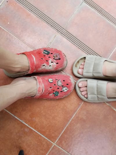 The image depicts two pairs of feet on a tiled floor. On the left, two feet are wearing red clogs adorned with various colorful charms, including a donut, a ghost, a daisy, and a leaf. The clogs appear worn and dusty. On the right, two feet are wearing light beige slides. The floor is composed of reddish-brown square tiles with gray grout lines. In the background, there are dark gray, ribbed strips, possibly part of a stairway or a transition strip. The overall scene is likely a domestic or outdoor area in Ban Patan, Thailand, given the context of the location. The image captures a static moment, with no discernible action or interaction between individuals.