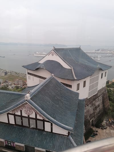An elevated, slightly hazy, eye-level view depicts the traditional architecture of a building, likely a temple or historical structure, in Hong Gai Ward, Vietnam. The building features multi-tiered, blue-tiled roofs and white walls. In the background, Halong Bay is visible, with numerous islands and boats, including a large cruise ship. A coastal city extends along the shoreline to the right. In the foreground, below the structure, the area appears to be under construction. Scooters are parked nearby, and construction material is scattered. The sky is overcast, suggesting a humid, coastal environment.