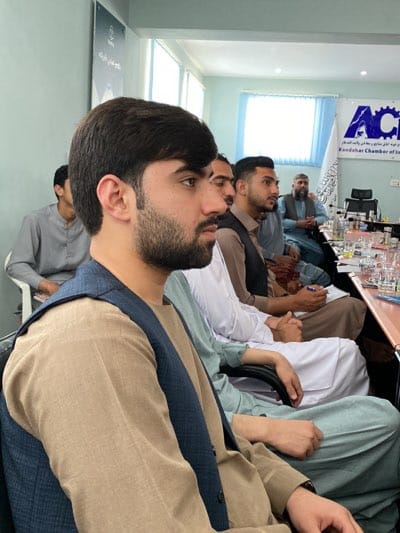 The image shows a meeting in what appears to be an office or conference room. Several men are seated around a table covered with water glasses and papers. The person closest to the camera is a young man with dark hair and a beard, wearing a light brown shirt and a dark blue vest. Behind him, other men are dressed in traditional Afghan clothing. In the background, there is a banner for the Kandahar Chamber of Industries and Mines (ACIM). The location is likely in Kandahar, Afghanistan, possibly in the Shurandam area, as that is the location of the Chamber of Industries and Mines.