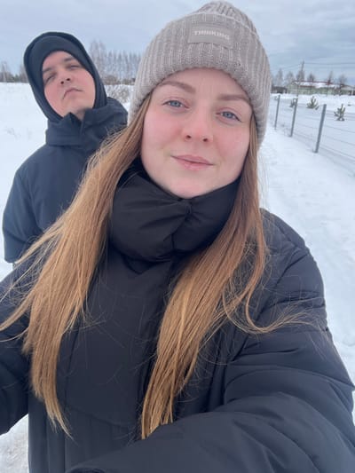 The image displays two individuals in a snowy outdoor environment in Kalinino, Russia, under an overcast sky. In the foreground, a female subject with light skin, blue eyes, and long, light brown hair is visible, wearing a light grey ribbed beanie with a rectangular "THINKING" patch and a black puffer jacket with a prominent high collar. She is positioned to face the camera, exhibiting a slight smile. Behind her and to her left, a male subject with light skin is partially visible, wearing a black hooded jacket with the hood up, and looking upwards towards the camera with a neutral expression. The ground surface is uniformly covered with a layer of white snow. The background features a snow-covered field with bare trees on the left side, while the right side shows a wire mesh fence running alongside a snowy path. Beyond the fence, several small evergreen shrubs are planted in a row, and distant light-colored houses are discernible under the cloudy sky.