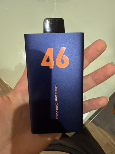 A close-up shot shows a dark blue vape pen with orange lettering that reads "46 MANGO PEACH". The vape pen is held in a person's left hand, with fingers visible on the right side of the frame. The background appears to be a light-colored, smooth surface, possibly a desk or table. The image is devoid of any urban context or recognizable landmarks that would allow for geolocation to a specific city like ЖК «Борисоглебское», Russia.