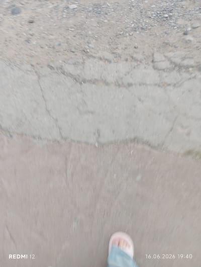 The image shows a person's foot wearing a light pink sandal and blue jeans, standing on a cracked concrete surface. To the left of the foot, the text "REDMI 12" is visible. To the right of the foot, the date "16.06.2026" and time "19:40" are displayed. The background consists of a mixture of dirt and small stones, suggesting an outdoor environment. The scene appears to be a snapshot taken while walking, as indicated by the motion blur. Based on the provided text, the image was captured in Bekabad, Uzbekistan.