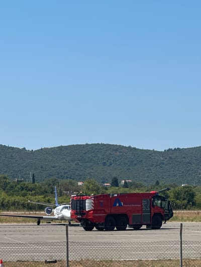 In Mrčevac, Montenegro, a red fire truck with the logo "Airports of Montenegro" is parked on an airport tarmac. To the left of the fire truck is a white and blue private jet. In the background, a lush green hillside with scattered houses is visible under a clear blue sky. The scene appears to be on a sunny day.