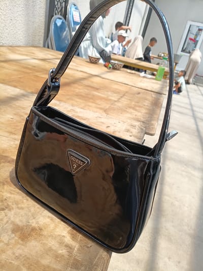 A black, patent leather Guess handbag with a triangular metal logo is placed on a wooden table. The handbag is in the foreground and is the main subject of the image. In the background, several people, including children and adults, are gathered around tables. The setting appears to be an outdoor cafe or market area in Osh, Kyrgyzstan, with some chairs and what looks like a building facade visible. The lighting suggests it is daytime.