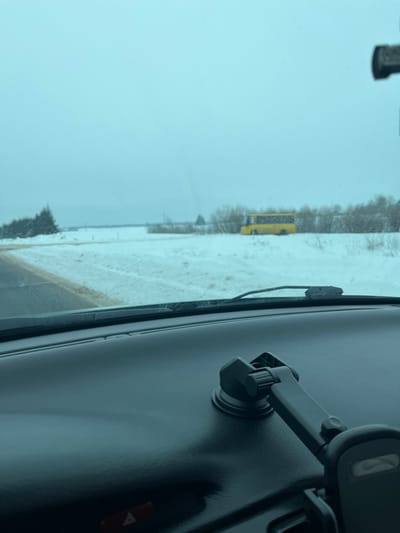 The scene depicts a winter landscape in Карабоўскі сельскі Савет, Belarus, viewed from inside a vehicle. The foreground shows a dark grey or black dashboard occupying the lower portion of the image. A black phone mount with a suction cup base is affixed to the dashboard on the right side, extending its arm towards the right. A red triangular hazard light button is visible on the lower left of the dashboard. A windshield wiper blade rests at the base of the windshield.
Beyond the windshield, a snow-covered environment is visible under an overcast, light grey sky. A two-lane asphalt road, partially covered with slush or dirt, runs along the left side of the frame, receding into the distance. The ground adjacent to the road and extending across the midground is covered in snow. A yellow bus is parked or stationary in the snow-covered field on the right side of the road, oriented with its side visible. Sparse, bare vegetation, including bushes and small trees, is scattered across the snowy field behind the bus. A dense cluster of dark green coniferous trees stands on the far left horizon. The overall impression is a cold, cloudy winter day. No individuals are visible.