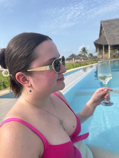 The image displays an adult female from the mid-torso up, partially submerged in a swimming pool under a clear blue sky. She wears a bright pink bikini top, gold-framed round sunglasses with dark lenses, a silver necklace featuring a key-shaped pendant, and a silver hook earring with a small, faceted blue stone on her visible left ear. Her dark hair is pulled back into a bun, secured by a large, clear, ornate hair clip. She holds a clear, stemmed glass containing a pale yellow liquid with condensation on its exterior in her raised right hand, while looking towards the right of the frame. The background shows green landscaping, a prominent building with a large, steeply pitched, dark thatched roof, and several smaller thatched structures and palm trees, indicative of a resort environment on the island of Zanzibar, Tanzania.