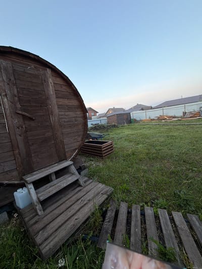 The image, originally oriented in portrait mode and rotated 90 degrees counter-clockwise, depicts a rural or suburban backyard scene in Большая Шильна, Russia. The sky, visible across the top and left, is clear and light blue, indicating daytime, likely early morning or late afternoon due to the soft, diffused light and absence of harsh shadows.
In the foreground, a dark brown, barrel-shaped wooden structure, possibly a shed or a small outbuilding, occupies the lower-left portion of the frame. Its rounded roof and vertical wooden planks are clearly visible. Next to it, on the right, a wooden panel or gate lies flat on the ground, and beneath it, a white plastic container is partially visible. Further to the right, a makeshift pathway of weathered wooden planks extends into the grassy area. A faint reflection, possibly of a person's face or hand from a smartphone screen, is visible on the far right edge of the image.
The midground features a patch of unkempt green grass with some weeds. Towards the center-right, a wooden slatted crate or planter box sits on the ground, with a blue object (possibly a wheelbarrow handle) partially seen behind it. In the background, a light-colored metal fence separates the yard from a residential area. Beyond the fence, several houses with dark roofs are visible, and a pile of stacked wooden planks or building materials can be seen near the fence.
No specific activity is taking place; the scene is static. There are no clearly visible individuals, apart from the possible reflection. No discernible text is present in the image.