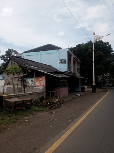 The image depicts an urban street scene in Chhenangri_Rishor, Bhutan, under an overcast sky. In the foreground on the left, a single-story, dilapidated structure with a dark, corrugated metal roof is visible. Its walls are partially exposed brickwork, and a faded banner displaying "SATE LINTAS" and an image of a woman hangs on its front. A small, green-leafed tree stands near this structure. Directly behind and slightly to the right of the foreground structure is a two-story building with a light blue upper facade and a dark tiled roof. It features a window with a metal security grille and an open balcony section.
The foreground consists of a dirt and gravel shoulder. On this shoulder, a rustic wooden table with a metal frame is positioned on the left, and a pile of cut wooden branches lies further to the right. Under the roof of the foreground structure, two stalls or display cabinets are visible, one appearing reddish-brown. To the right, a paved asphalt road runs parallel to the dirt shoulder, marked by a continuous yellow line on its edge and curving gently to the right in the distance. A tall, white streetlamp post with decorative gold-colored scrollwork at its top stands on the right side of the dirt shoulder, featuring a single light fixture extending horizontally. Dense green trees are visible in the background, primarily behind the buildings on the right. No individuals or active interactions are observed.