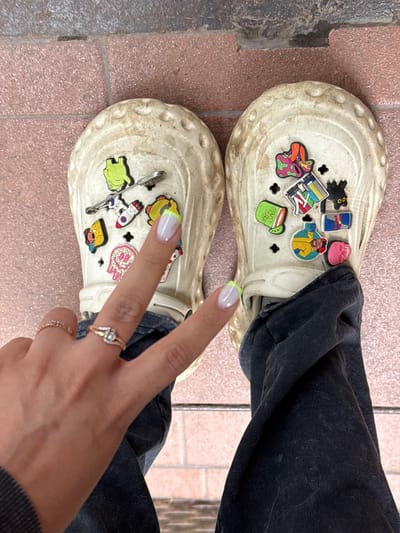 The image shows a close-up view of a person's feet wearing beige Crocs adorned with various colorful charms. The Crocs are placed on a tiled surface, with some stonework visible to the left. The person is wearing dark denim pants.
The charms on the left Croc include a green frog, a rocket ship, and a safety pin. The right Croc features a black cat, a Red Bull can, a pink skull, a person in a yellow jacket, a cocktail, and a colorful "MUSIC TELEVISION" charm. A hand with a manicure is visible in the lower right corner, making a peace sign and wearing two rings, one with a heart-shaped diamond.
The lighting suggests it is daytime, and the overall scene appears casual, possibly outdoors given the stonework. No specific time of day or weather conditions can be determined, and the location context of Naro-Fominsk, Russia, is provided but not visually evident in the image itself. The mood conveyed is playful and personalized due to the decorative charms on the footwear.