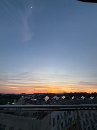 The image shows a vertical view of a sunset sky transitioning from dark blue to orange. Wispy clouds streak horizontally across the gradient. A crescent moon is visible in the upper left. On the right edge, a cityscape is visible, including buildings, structures with square panels, and what appears to be dense foliage below. The buildings are constructed of concrete. The image is slightly rotated.